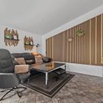 Wohnzimmer mit Sofa, Sessel und Holzwand. Fenster, Teppich und Couchtisch sichtbar.