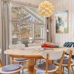 Round wooden table with chairs, garden view through window, lamp and picture on wall.