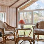 Two rattan chairs with fur covers stand by large windows. Between them is a small table with a lamp.