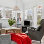 Living room with sofa, armchair, coffee table, and TV. Large windows and white walls.