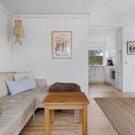 Living room with sofa, table, and kitchen in background. White walls and wooden floor.