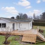 House with wooden terrace and garden. White facade, stairs, and seating area.