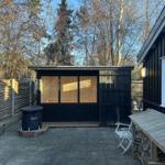 Backyard with sauna, wooden bench, folding chair, and woodpile. Surrounded by trees and log fence.
