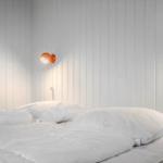 Schlafzimmer mit weißem Bett und orangefarbener Wandlampe an vertikal verkleideter Wand.