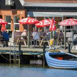 Hafenrestaurant mit Tischen unter roten Sonnenschirmen und blauem Boot im Wasser.