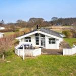 A white holiday home with a terrace and garden. Trees and a lake are visible in the background.
