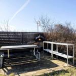 Outdoor area with table, benches, and wood stove. Background: fence and shrubs.