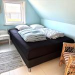 Schlafzimmer mit Bett, Nachttisch und Stuhl. Wandfarbe hellblau, Fenster mit Blick nach draußen.