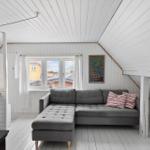 Wohnraum mit grauem Sofa, Fenster und Wandbild. Weiße Holzwände und Decke.