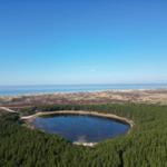Ein See umgeben von Wald, mit Dünen und Meer im Hintergrund.
