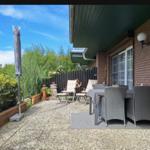 Terrasse mit Tisch, Stühlen und Sonnenschirm. Hintergrund: grüne Pflanzen und Zäunung.