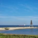 Hafen mit Segelboot, Sandstrand und Windkraftanlagen im Hintergrund.