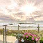 Terrasse mit Blick auf Strand und Meer bei Sonnenuntergang. Blumen und Glasgeländer sind sichtbar.