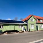Grünes Haus mit Solaranlage und rotem Dach. Straße und kleine Bäume sind sichtbar.