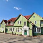 Grünes Holzhaus mit rotem Dach und weißen Fensterrahmen an sonnigem Tag.