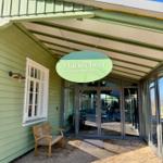 Eingangsbereich mit Schild, Glasfront und Holzbank vor grünem Haus