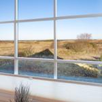 Großes Fenster mit Blick auf grasbewachsene Dünen und Horizont.