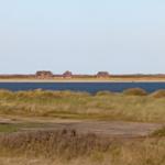Drei Häuser am Strand mit Dünen im Vordergrund und Wasser im Hintergrund.