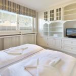 Schlafzimmer mit Doppelbett, weißen Schränken und TV. Fenster mit Rollladen und Blick auf Dächer.