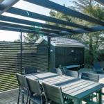 Terrasse mit Tisch, Stühlen und Sonnenschutz unter Pergola. Hintergrund: Wald und kleines Gebäude.