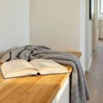 Ein offenes Buch liegt auf einer Holzbank mit einer Decke. Im Hintergrund ist ein Flur mit Holzboden.