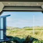 Durch ein großes Fenster blickt man auf Dünen und ein modernes Haus.