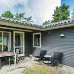 Haus mit Terrasse, Sitzgelegenheiten und Holzschild mit Namen. Umgeben von Bäumen und blauem Himmel.