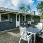 Ein graues Ferienhaus mit Terrasse und Gartenzaun. Gartenmöbel stehen auf dem gepflasterten Hof.