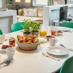 Rundtisch mit Frühstück, grüne Stühle, Wohnbereich im Hintergrund.