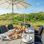 Terrasse mit Tisch, Stühlen und Sonnenschirm. Frühstückszubehör und Blick auf Grünflächen.