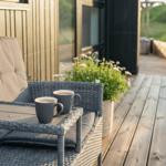 Terrasse mit Stuhl, Tisch und zwei Tassen Kaffee. Hintergrund: Grünflächen und Holzhaus.