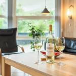 Holztisch mit Weinglas und Flasche, schwarze Ledermöbel, Fenster mit Blick auf Grün.