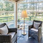 Zwei Rattan-Stühle mit Kissen stehen vor großen Fenstern mit Blick auf eine Terrasse und Wald.