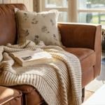 Ein braunes Sofa mit Kissen, Decke und Buch. Im Hintergrund steht ein Kaminofen.