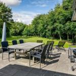 Terrasse mit Tisch, Stühlen und Grill neben Haus. Grünflächen und Bäume im Hintergrund.