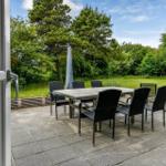 Terrasse mit Tisch, Stühlen und Grill. Grünfläche und Bäume im Hintergrund.