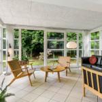 Ein Wohnzimmer mit Sofa, Stühlen und Fernseher. Große Fenster zeigen einen Garten.