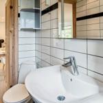Badezimmer mit Waschbecken, Toilette und Spiegel. Durchgang ins Wohnzimmer mit Tisch und Stühlen.