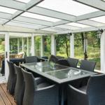 Terrasse mit Glasdach, Esstisch und Stühlen. Hintergrund: grüne Bäume und Schiebetüren.