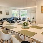 Essbereich mit Tisch und Stühlen. Im Hintergrund Wohnbereich mit Sofa und Fenstern.