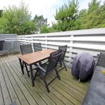 Terrasse mit Tisch, Stühlen, Grill und Liegestühlen. Hintergrund: Holzgeländer und Bäume.