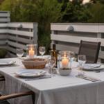 Tisch mit Tischdecke, Gläsern, Kerzen und Besteck auf einer Terrasse mit Sitzgelegenheiten.