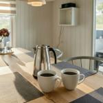 Esstisch mit Kaffeekanne und Tassen. Draußen ist ein Gartenbereich mit Tisch und Stühlen.