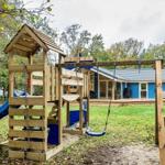 Holzspielplatz mit Rutsche und Schaukel vor einem blauen Haus mit Terrasse.