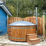 Holzwhirlpool mit Metallrahmen neben blauem Gebäude im Wald.