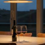 Ein Tisch mit Weinflasche und zwei Gläsern vor großen Fenstern.