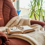 Ein braunes Sofa mit einer Decke und einem Buch. Im Hintergrund steht eine Pflanze.