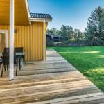 Holzterrasse mit Tisch und Stühlen neben gelbem Haus. Grasfläche mit Schaukel im Hintergrund.