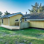 Ein gelbes Ferienhaus mit Holzveranda und Garten. Umgeben von Bäumen und Gras.