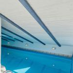 Innere Schwimmhalle mit blauem Wasser und Holzbalken an der Decke.